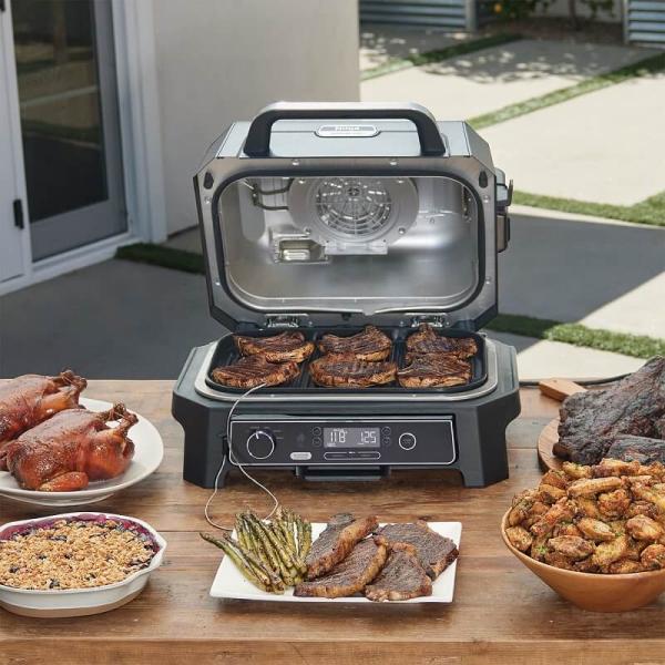A stylish and modern outdoor cooking setup featuring the Ninja Woodfire Grill in use, with woodfire smoke enhancing the scene. The grill is shown grilling meat, vegetables, and other dishes on a cozy patio surrounded by wooden furniture and potted plants. The image highlights the versatility of the Ninja Woodfire Grill and includes an overlay text that reads "Save 20% with Code NINJA20," emphasizing the discount available. The atmosphere is warm and inviting, showcasing the grill as the centerpiece of a perfect outdoor cooking experience.